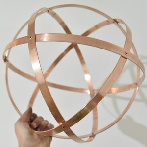 May include: A copper-colored metal sphere composed of intersecting bands. The bands are riveted together at the points of intersection, forming a three-dimensional geometric shape. The sphere is held by a hand against a white background.