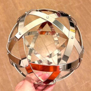 May include: A decorative silver metal sphere with intersecting bands, secured by small rivets. The reflective orb has a geometric design, suitable for home decor.