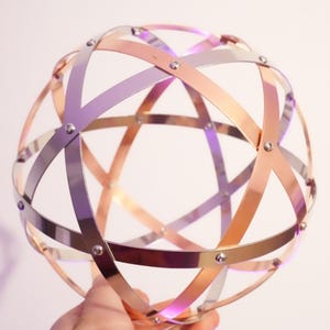 Può includere: Una sfera decorativa in metallo con fasce intersecate nei toni del rame, argento e viola. Le fasce sono rivettate insieme, formando un motivo geometrico. La sfera è tenuta in mano su sfondo bianco.