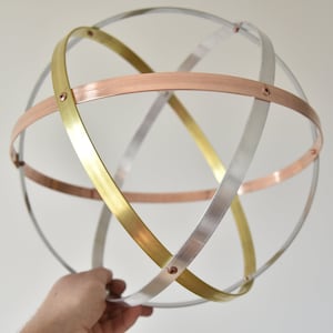 May include: A decorative metal sphere with intersecting bands in silver, gold, and copper. The bands are riveted together, forming a geometric pattern. The sphere is held by a hand. The sphere's diameter is approximately 15 cm.