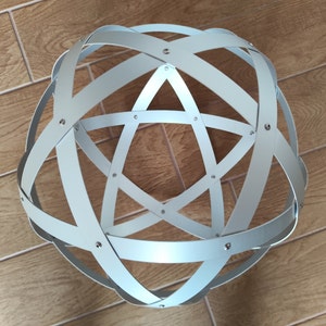 Può includere: Una scultura sferica in metallo grigio chiaro con fasce intersecanti che formano un motivo geometrico. Le fasce sono fissate con piccoli rivetti, creando un oggetto decorativo moderno. Un'opera d'arte unica.