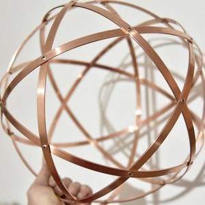 May include: A copper sphere sculpture made from intersecting metal bands, riveted at the intersections. The geometric design is held against a white background, creating a shadow. The sphere's diameter is approximately 20 cm.