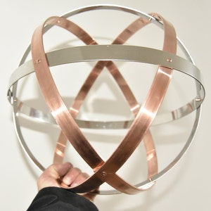 Può includere: Una scultura sferica con fasce intersecanti di rame e metallo color argento. Le fasce sono disposte per creare un design geometrico tridimensionale. La scultura è tenuta da una persona.