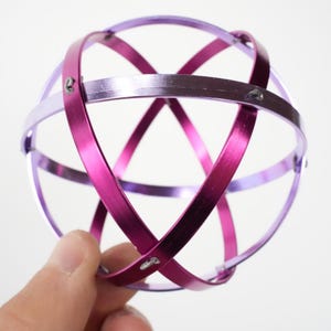 Op de afbeelding: Een geometrische, bolvormige sculptuur bestaande uit elkaar kruisende banden. De banden zijn helder magenta en een lichtere lavendelkleur, waardoor een driedimensionale bol ontstaat. Kleine metalen bevestigingen houden de banden bij elkaar.