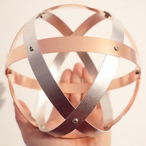 May include: A spherical decorative object made from intersecting copper and silver metal bands. The bands are riveted together, creating a cage-like design. The sphere is held in a hand, showing its size and construction.