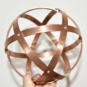 May include: A copper-coloured metal sphere made from intersecting curved bands. The bands are riveted together, creating a geometric, openwork design. The sphere is held in a hand against a white background.