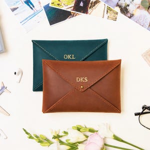 May include: Two envelope-style leather pouches, one in dark green and one in brown, featuring gold-embossed initials. The green pouch is monogrammed with OKL, while the brown pouch is monogrammed with DKS and secured with a small gold-toned metal snap.