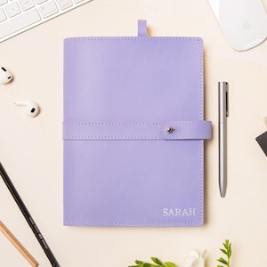 May include: A lavender leather journal with the name "SARAH" embossed on the cover, next to a silver pen, a white computer mouse, and a keyboard. The journal is closed with a strap and button. Pencils, an eraser, and a magazine are also present.
