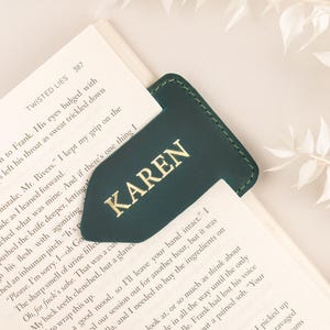 May include: Dark green leather bookmark with the name "KAREN" in gold lettering, inserted into a book. The bookmark has a unique arrow-like shape. The book is open to a page of text.