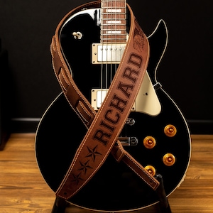 May include: A black electric guitar with a brown leather strap. The strap is embossed with the name "RICHARD" and star designs. The guitar has gold-colored knobs and is on a black stand.