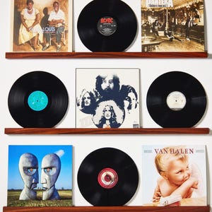 May include: A collection of vinyl records displayed on three wooden shelves. The album covers feature various artists and album art, including AC/DC, Van Halen, and Pink Floyd. The records are black with visible labels.
