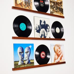Può includere: Un'esposizione a parete di dischi in vinile e copertine di album. I dischi sono neri con etichette colorate. Le copertine degli album presentano varie immagini e testi, montati su tre mensole di legno. I dischi sono esposti su una parete bianca.