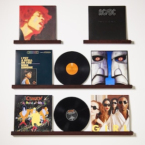 May include: A display of vinyl records on dark brown shelves against a white wall. The records showcase album art from artists including Jimi Hendrix, AC/DC, Nina Simone, and Queen. The records are black with orange and yellow labels.