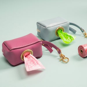 May include: Two small, rectangular pet waste bag dispensers in pink and blue, with gold-coloured hardware and matching straps. A roll of red bags with white text is next to them. Green and pink bags are visible.