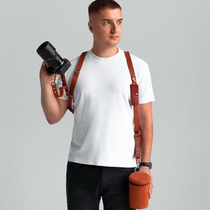 May include: A brown leather camera harness with a matching lens case. The harness has adjustable straps and metal hardware. The lens case is cylindrical and has a zip closure. The camera is black.