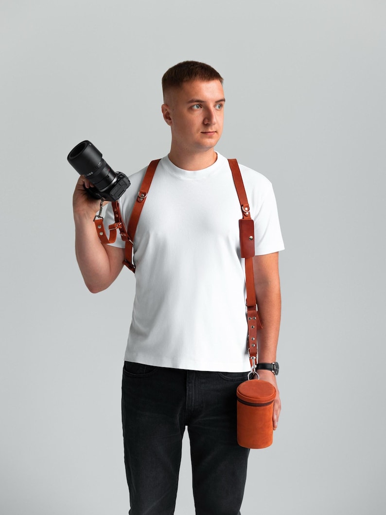 May include: A brown leather camera harness with a matching lens case. The harness has adjustable straps and metal hardware. The lens case is cylindrical and has a zip closure. The camera is black.