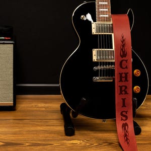 Puede incluir: Una guitarra el&eacute;ctrica negra con una correa de cuero roja. La correa est&aacute; personalizada con el nombre "CHRIS" y dise&ntilde;os de llamas. La guitarra est&aacute; sobre un soporte negro, con un amplificador Marshall y una c&aacute;mara en el fondo.