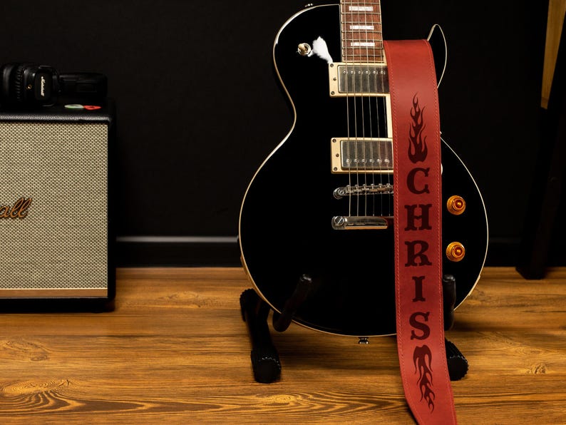 Puede incluir: Una guitarra el&eacute;ctrica negra con una correa de cuero roja. La correa est&aacute; personalizada con el nombre "CHRIS" y dise&ntilde;os de llamas. La guitarra est&aacute; sobre un soporte negro, con un amplificador Marshall y una c&aacute;mara en el fondo.