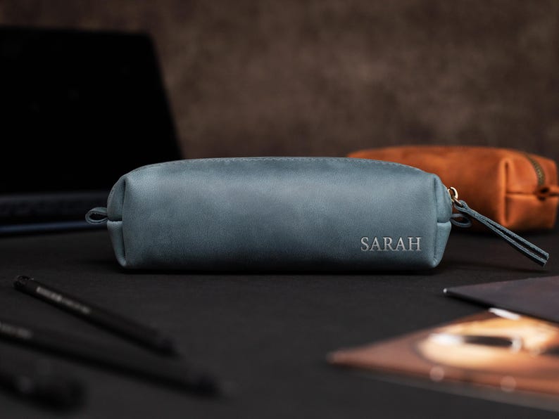 May include: A teal leather pencil case with the name "SARAH" embossed on the front. The case has a zipper closure and a small wrist strap. Another brown leather case is in the background. The items are on a black surface.