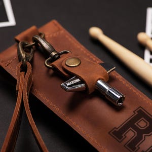 May include: A brown leather drum key holder with a metal drum key and a strap. The holder has a snap closure and a metal clasp. The word "MAXTONE" is visible on the drum key. Two wooden drumsticks are in the background.