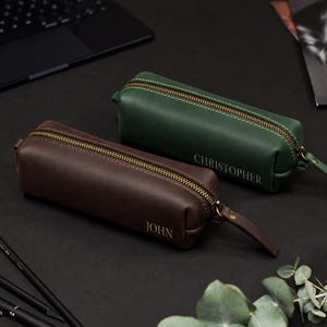 Pode incluir: Dois estojos retangulares de couro liso, um em castanho escuro com o nome JOHN gravado e outro em verde floresta com o nome CHRISTOPHER gravado. Ambos os estojos possuem um fecho de metal cor de latão e um puxador de couro a condizer.