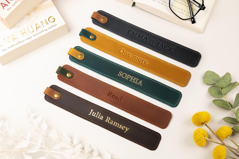 May include: Leather bookmarks in various colors, including black, mustard yellow, and brown. Each bookmark has a stitched border and a snap closure. Some bookmarks are personalized with names or phrases like "Emma & Jack" and "Read."