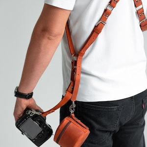 May include: A tan leather camera harness with a small, matching pouch. The harness features adjustable straps and metal fixings. The camera is black, and the pouch is a small, square shape with a zip.