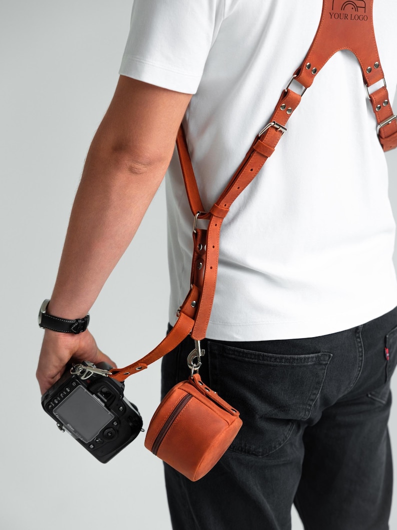May include: A tan leather camera harness with a small, matching pouch. The harness features adjustable straps and metal fixings. The camera is black, and the pouch is a small, square shape with a zip.