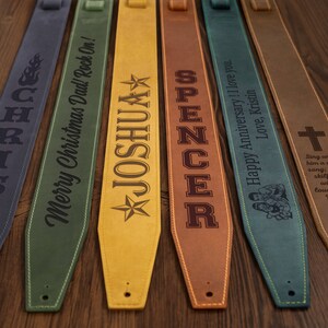 May include: A collection of personalized guitar straps in various colors, including blue, green, yellow, and brown. Each strap features custom text and designs, such as names, flames, and a cross, making them unique gifts.