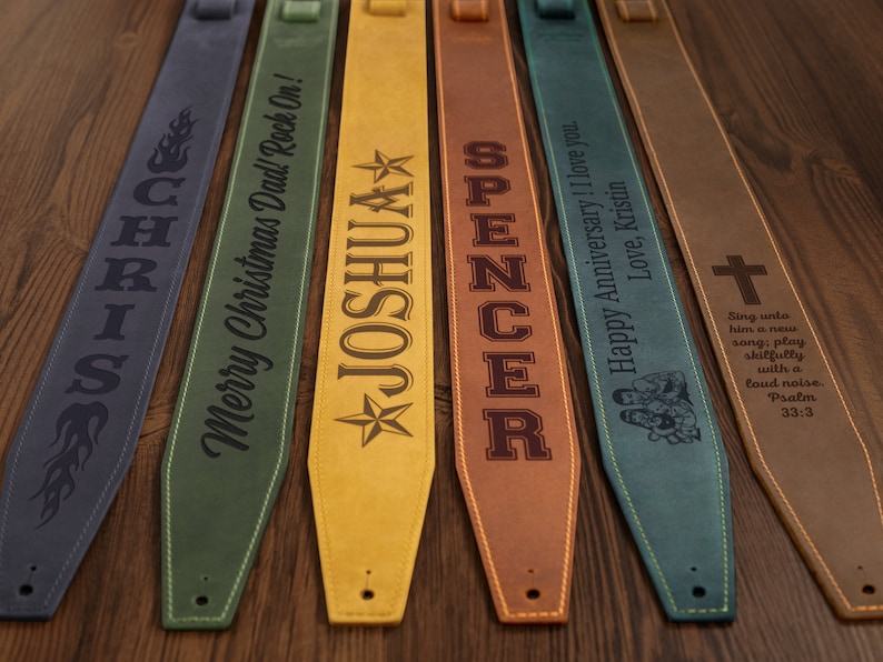 May include: A collection of personalized guitar straps in various colors, including blue, green, yellow, and brown. Each strap features custom text and designs, such as names, flames, and a cross, making them unique gifts.