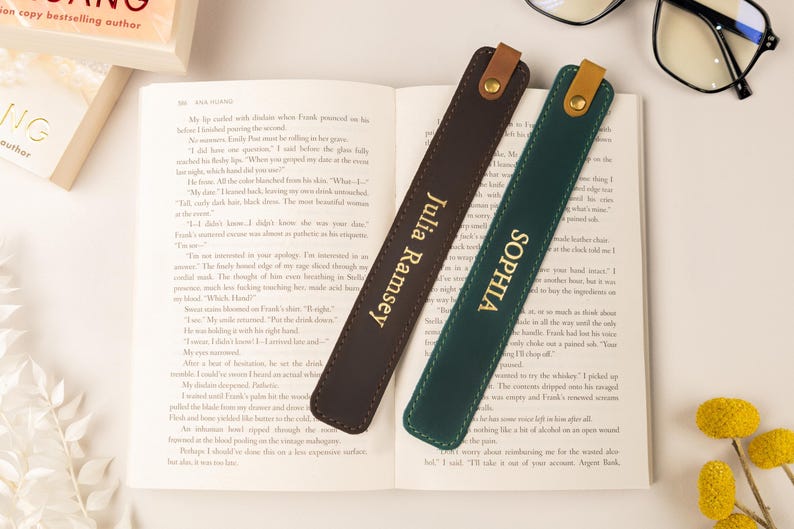 May include: Two leather bookmarks, one brown and one green, rest on an open book. The brown bookmark has "Julia Ramsey" in gold lettering, and the green one reads "Sophia". Each has a leather loop.