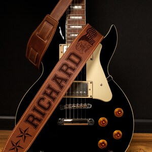 May include: A black electric guitar with a brown leather guitar strap. The strap is embossed with the name "RICHARD", an eagle, stars, and stripes. The guitar has gold knobs and a cream-colored pickguard.
