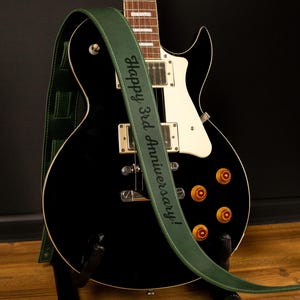 K&ouml;nnte beinhalten: Eine schwarze E-Gitarre mit einem gr&uuml;nen Lederriemen. Auf dem Riemen steht in wei&szlig;en Buchstaben "Happy 3rd Anniversary!". Die Gitarre hat goldene Kn&ouml;pfe und wei&szlig;e Akzente. Der Hintergrund ist dunkel.