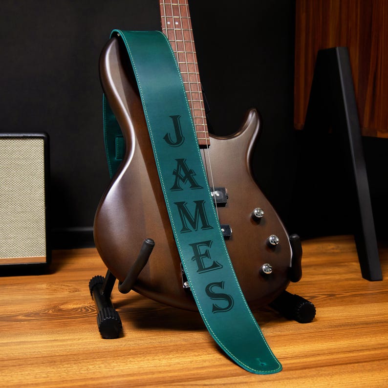 May include: A brown electric bass guitar with a green leather strap. The strap is embossed with the name "JAMES" in black capital letters. The guitar is on a black stand, with a small amplifier visible in the background.