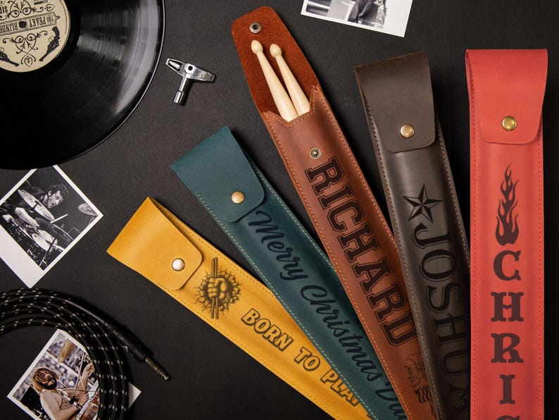 May include: A collection of personalized leather drumstick cases in various colors, including brown, green, and red. Each case features custom text like "RICHARD" and "JOSHUA", and some include graphics. Drumsticks are visible in some cases.