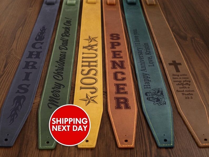 K&ouml;nnte beinhalten: Eine Auswahl an personalisierten Gitarrengurten in verschiedenen Farben, darunter Blau, Gr&uuml;n, Gelb und Braun. Jeder Gurt zeigt individuellen Text und Designs, wie Namen, Phrasen und ein Kreuz. Die Gurte sind aus leder&auml;hnlichem Material gefertigt.