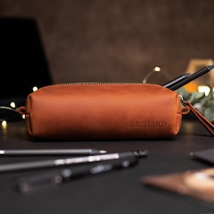 May include: A brown leather pencil case with a gold zipper and the name "RICHARD" embossed on the front. The case is cylindrical and contains a black pen. Other pens are in the foreground.
