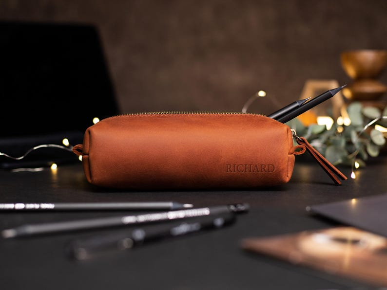 May include: A brown leather pencil case with a gold zipper and the name "RICHARD" embossed on the front. The case is cylindrical and contains a black pen. Other pens are in the foreground.