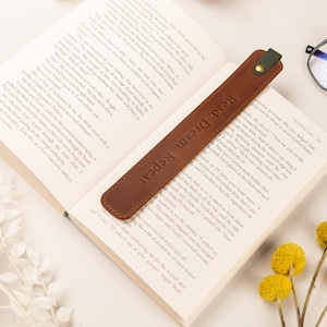 May include: A brown leather bookmark with the words "Read. Dream. Repeat." embossed on it, resting on the pages of an open book. The bookmark has a green accent and a gold button. A pair of glasses and yellow flowers are also in the image.