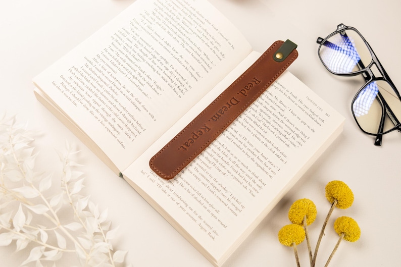 May include: A brown leather bookmark with the words "Read. Dream. Repeat." embossed on it, resting on the pages of an open book. The bookmark has a green accent and a gold button. A pair of glasses and yellow flowers are also in the image.