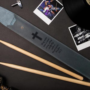 May include: A gray leather drumstick case with a black cross and text, accompanied by two wooden drumsticks. The case is open, revealing its interior. The image includes a microphone, photos, and a vinyl record.