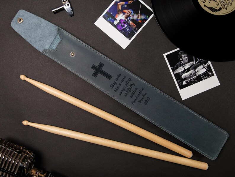 May include: A gray leather drumstick case with a black cross and text, accompanied by two wooden drumsticks. The case is open, revealing its interior. The image includes a microphone, photos, and a vinyl record.