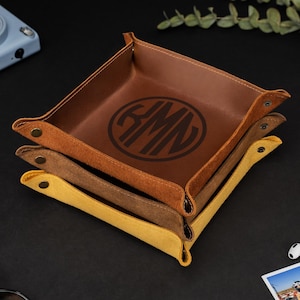 Könnte beinhalten: Ein Stapel von drei Leder-Ablageschalen in Braun- und Gelbtönen. Die obere Schale ist ein sattes Braun mit einem Monogramm. Die Schalen sind quadratisch mit abgerundeten Ecken und mit Metallschnappern befestigt. Zubehör ist verstreut.