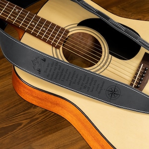 Puede incluir: Guitarra ac&uacute;stica con una correa de guitarra de cuero gris oscuro. La correa tiene costuras blancas y texto grabado con un dise&ntilde;o de rosa de los vientos. La guitarra descansa sobre una superficie de madera.