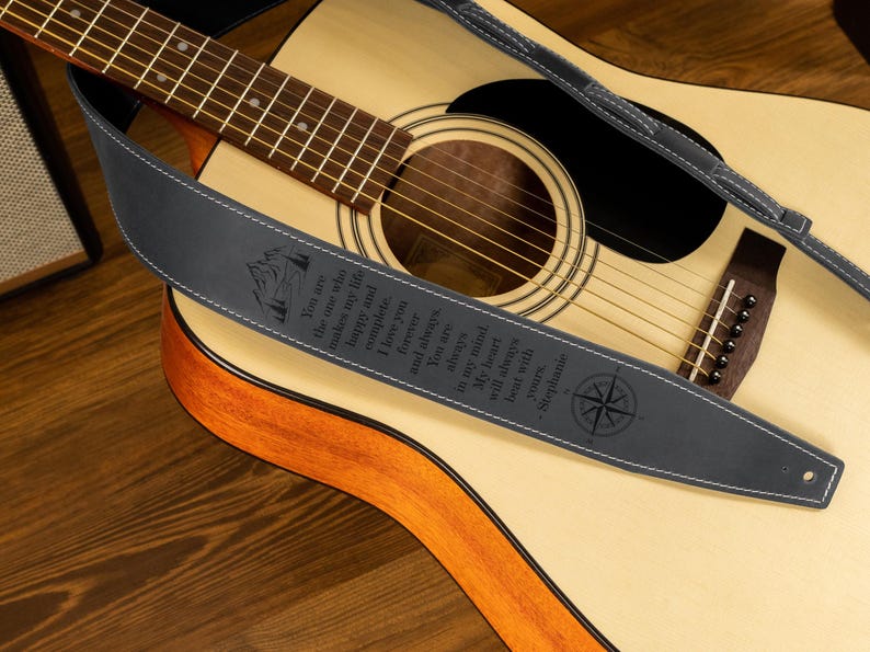 Puede incluir: Guitarra ac&uacute;stica con una correa de guitarra de cuero gris oscuro. La correa tiene costuras blancas y texto grabado con un dise&ntilde;o de rosa de los vientos. La guitarra descansa sobre una superficie de madera.