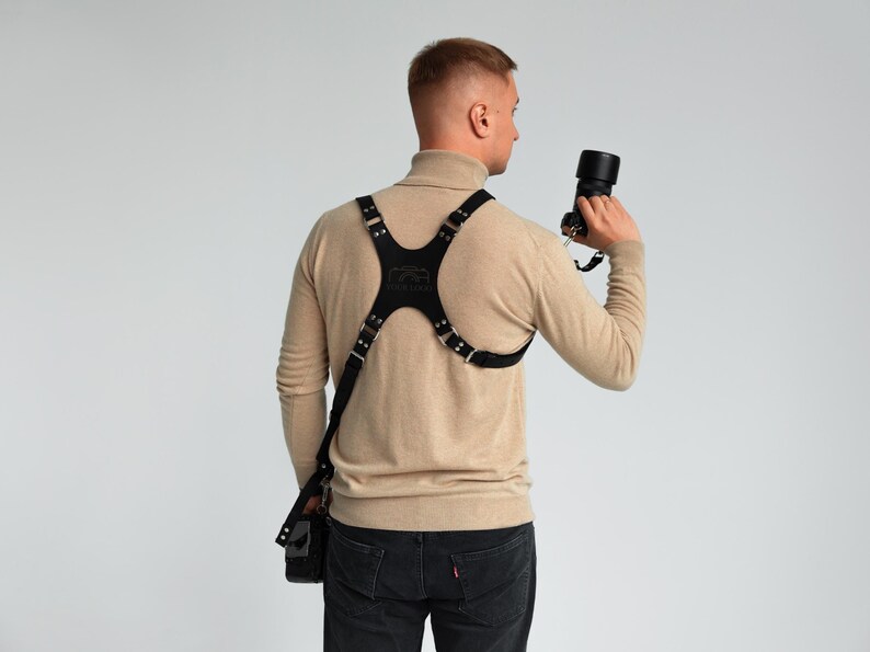May include: Black leather camera harness with adjustable straps and a chest plate. The harness is designed to hold a camera and lens, and it has a small pouch. The person is holding a black camera with a lens.