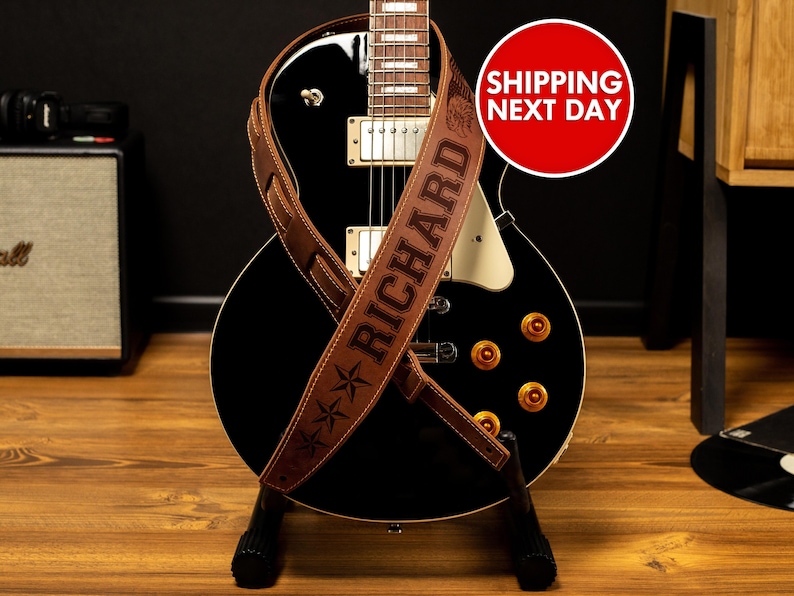 Puede incluir: Una correa de guitarra de cuero marr&oacute;n con el nombre "RICHARD" grabado y dise&ntilde;os de estrellas, sobre una guitarra el&eacute;ctrica negra. La guitarra tiene perillas doradas y un golpeador blanco. Tambi&eacute;n se ve un c&iacute;rculo rojo con texto blanco.