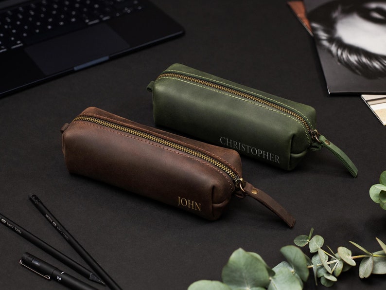 May include: Two leather pencil cases, one brown and one green, with brass zippers and personalized names. The brown case is embossed with "JOHN", and the green case with "CHRISTOPHER". Both cases have a wrist strap.