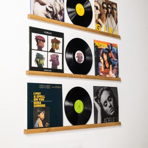 May include: A display of vinyl records on wooden shelves against a white wall. The records include album covers from Gorillaz, Nina Simone, and Adele. The records are black with colorful labels.