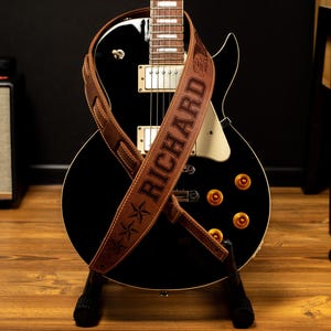 K&ouml;nnte beinhalten: Eine schwarze E-Gitarre mit einem braunen Lederriemen. Der Riemen ist mit dem Namen "RICHARD" und Sternmotiven gepr&auml;gt. Die Gitarre hat goldfarbene Kn&ouml;pfe und steht auf einem schwarzen St&auml;nder.